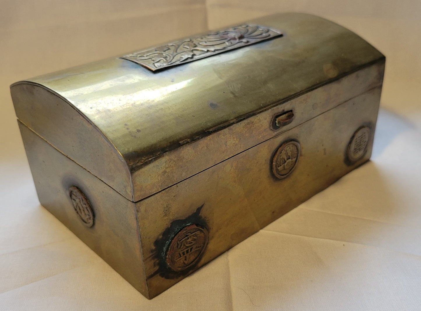 Antique brass box curio made in China, with a domed lid. The interior bottom has wood inserts on three side (one side is missing). The exterior has welded medallions with Chinese characters and designs. View of corner angle.